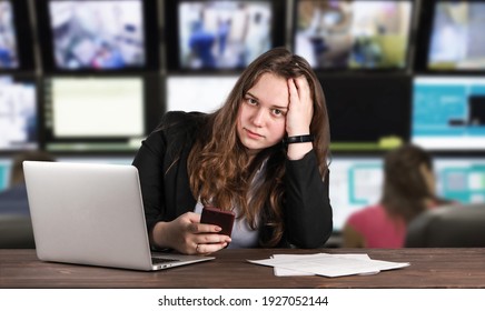 Portrait of young woman sitting at her workplace with silver laptop on her table. She wears black strict formal suit. Lots of TV broadcasting screens in the background. Lady looks tired and fatigued.