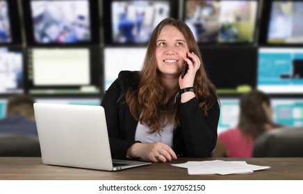 Porträt von jungen Frauen, die auf ihrem Arbeitsplatz mit silbernem Laptop auf ihrem Tisch sitzen. Sie trägt einen schwarzen, strengen formalen Anzug. Viele Fernsehbildschirme im Hintergrund. Sie telefoniert gerade.