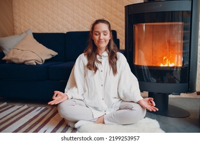 Portrait of young woman sitting in lotus position indoors with fireplace. Yoga and meditation zen like concept
