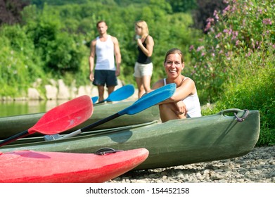 Portrait Of A Young Woman Sitting Next To A Kayak On A Riverbank, Friends In The Background