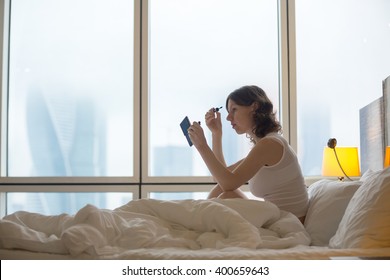 Portrait Of Young Woman Sitting On Unmade Bed After Waking Up And Putting On Mascara. Caucasian Female Model Getting Ready For Working Day At Home. Copy Space