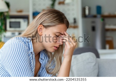 Portrait of a young woman sitting on the couch at home with a headache and pain. Beautiful woman suffering from chronic daily headaches. Sad woman holding her head because sinus pain