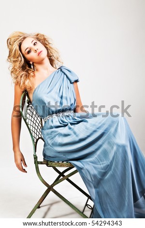 Portrait of a young woman sitting on a chair. Her hair is wild and she is wearing a blue dress which is off one shoulder. Horizontal shot. Isolated on white.