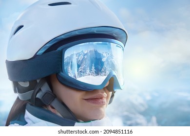 Portrait Of Young Woman At The Ski Resort On The Background Of Mountains And Blue Sky.A Mountain Range Reflected In The Ski Goggles. Winter Sports.