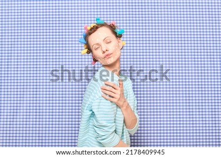 Portrait of young woman with skincare product on face, with colorful hair curlers on head, having fun, fooling around, pouting lips, making face, standing in bathroom over shower curtain background