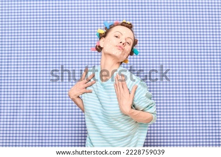 Portrait of young woman with skincare product on face, with colorful hair curlers on head, having fun, fooling around, pouting lips, making face, standing in bathroom over shower curtain background