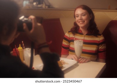 Portrait of young woman smiling while sitting in retro diner booth, red-striped sweater and milkshake on table, photographer capturing moment - Powered by Shutterstock