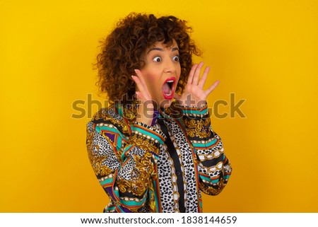 Portrait of young woman standing against yellow background, with shocked facial expression holding hands near face, screaming and looking sideways at something amazing.