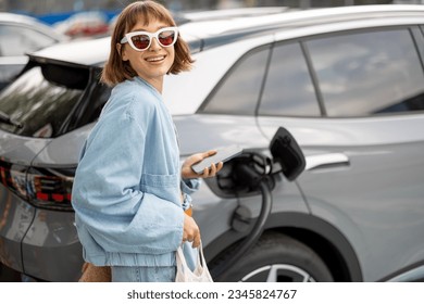 Portrait of a young woman stands with a smart phone, waiting for her electric car to charge on a public charging station outdoors. Concept of modern lifestyle and green energy for transportation - Powered by Shutterstock