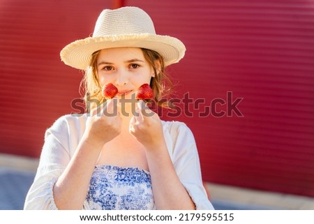 Portrait of young woman in staw hat tasting a fresh strawberry on red backgtound outdoor. Summer mood.
