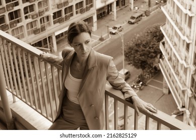 Portrait Of Young Woman In Suit Standing On Balcony With Busy City Street In Background	
