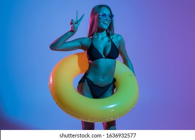 Portrait of young woman with sunglasses in bikini playing with yellow inflatable ring on blue background