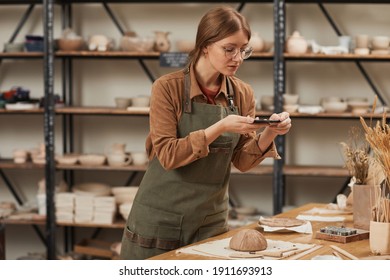 Portrait of young woman taking smartphone photo of raw ceramic bowl in pottery workshop to post on social media, small business concept, copy space - Powered by Shutterstock