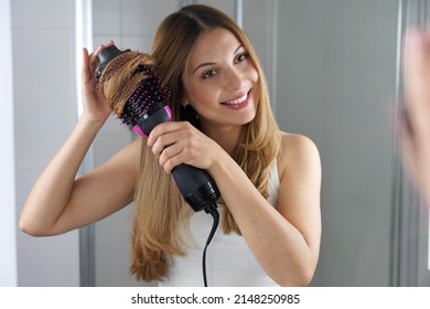 Portrait of young woman using round brush hair dryer to style hair at the mirror in an easy way at home - Powered by Shutterstock