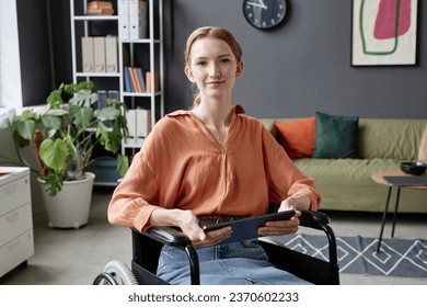 Portrait of young woman using wheelchair smiling at camera and holding tablet in office, copy space - Powered by Shutterstock