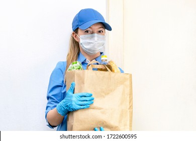 Portrait Of A Young Woman Volunteer In A Blue Uniform And Cap, In A Medical Mask And Gloves Holds A Paper Bag With Food And Vegetables In Her Hands. Donation, Help People In Quarantine, Coronavirus.