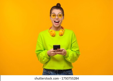 Portrait of young woman wearing colored eyeglasses, green neon sweatshirt and headphones around neck, excited by content on phone screen, isolated on yellow background - Powered by Shutterstock