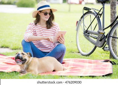 Portrait of a young woman wearing straw hat and sunglasses while sitting at park with her puppy and using digital tablet.  - Powered by Shutterstock