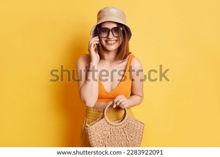 Portrait of young woman wearing summer clothing with straw bag standing isolated over yellow background talking on smart phone, having pleasant conversation
