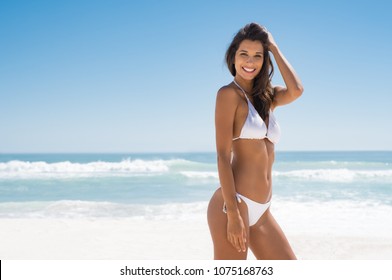 Portrait of young woman in white bikini on tropical beach looking at camera. Beautiful latin girl in swimwear with copy space. Summer vacation and tanning concept.