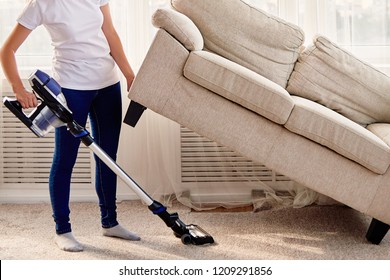 Portrait of young woman in white shirt and jeans cleaning carpet under sofa with vacuum cleaner in living room, copy space. Housework, cleanig and chores concept