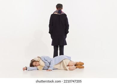 Portrait Of Young Woman In Winter Coat Lying On Floor And Sleeping, Man Standing Behind Her Over White Background. Concept Of Retro Fashion, Style, Friendship, Creativity, Youth Culture, Emotions, Ad