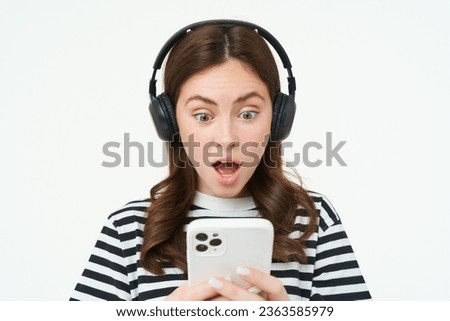 Portrait of young woman in wireless headphones, looks shocked at smartphone screen, reads message on mobile phone with dropped jaw, gasping from surprise, white background.