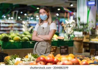 Portrait Young Woman Worker Seller In A Vegetable Section Supermarket Standing In A Protected Face Mask Arms Crossed. Greengrocer Female Looking At Camera In Fruit Shop Market Employee In A Work Apron