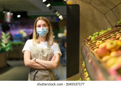 Portrait Young Woman Worker Seller In A Vegetable Section Supermarket Standing In A Protected Face Mask Arms Crossed. Greengrocer Female Looking At Camera In Fruit Shop Market Employee In A Work Apron