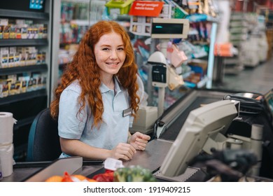 Portrait Of A Young Woman Working At Grocery Store Checkout Counter. Female Cashier Sitting Behind Checkout Counter Looking At Camera And Smiling.