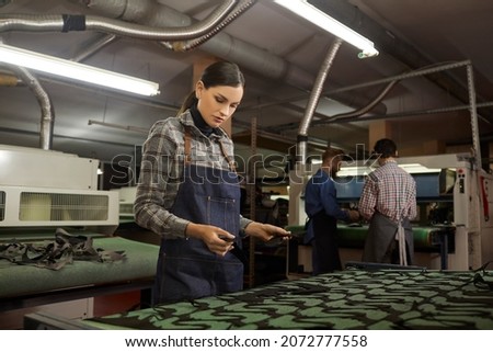 Portrait of young woman working in workshop of footwear factory. Serious female worker standing at cutting machine and looking at cut details of leather material. Concept of shoe manufacturing process
