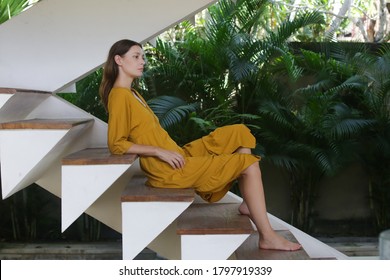 Portrait Of Young Woman In Yellow Dress Sitting At Home On The Wooden Stairs, Side View