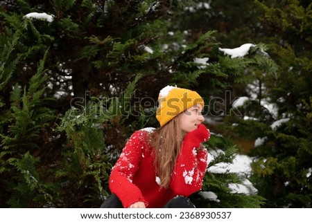 Portrait of a young woman in a yellow hat covered in snow having fun outdoors against a background of Christmas trees. Concept of winter activities and entertainment, celebration of the new year