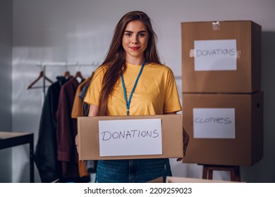 Portrait Of A Young Woman In A Yellow T Shirt Holding Cardboard Box With Donation Label While Working In A Charity Foundation. Humanitarian Aid Concept. Female Working In Homeless Shelter.