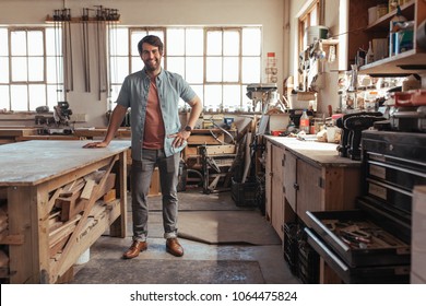 Portrait Of A Young Woodworker Smiling Confidently While Standing Next To A Workbench Full Of Wood In His Carpentry Workshop 