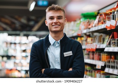 Portrait Of A Young Worker At A Grocery Store And Looking At The Camera Smiling. Man In Uniform Standing With Arms Crossed In Supermarket.