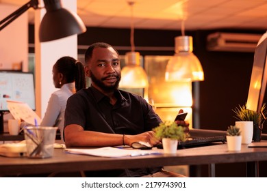 Portrait Of Young Worker Using Smartphone App At Office Job, Working With Mobile Phone And Computer To Create Business Report. Browsing Social Media Network To Do Paperwork Research.