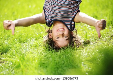 Portraits Of Happy Kid Playing Upside Down Outdoors In Summer Park With Thumbs Up