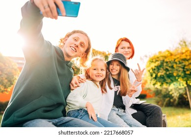 Portraits Of Three Smiling Sisters And Brother Teen Taking Selfie Portrait Using A Modern Smartphone Camera. Careless Young Teenhood And Childhood Time And Modern Technology Concept Image.