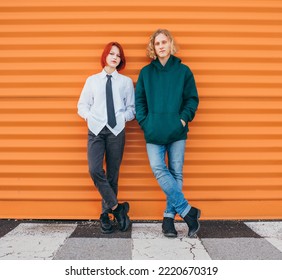 Portraits Of Two Caucasian Teen Friends Boy And Girl Posing For Photo While They Standing Near The Orange Wall Background. Careless Young Teenhood. Time Concept Image.