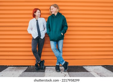 Portraits Of Two Smiling To Each Other Caucasian Teen Friends Boy And Girl Casual Dressed While They Standing Lean On The Orange Wall Background. Careless Young Teenhood Time And First Love Concept.