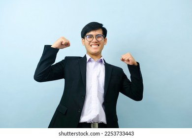 Portriat of asian young smart happy businessman wearing glasses and dressed in suit standing smiling, and flexing at camera to show strength and powerful positive expression with isolated light blue.