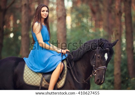 Portriat of smiling young beautiful brunette girl in blue dress ride on the black horse in forest