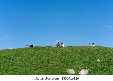 Portsmouth, Hampshire, UK - 24 March 2019: Three Couples Sit On Top Of A Grassy Hill Enjoying The Sunshine Under A Clear Blue Sky. Men, Women And A Child Sit In Pairs Maintaining Personal Space.