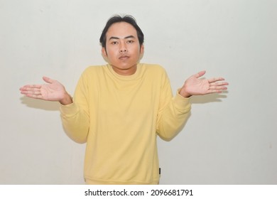 Pose Of Asian Man Shrugging Both Shoulders And Raising Both Hands. Illustrations Of Ignorance, Why Not, Where, And What's Wrong. Portrait Of Indonesian Man On Isolated White Background	
