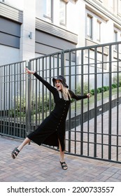 Posing Blond Woman Covered In Black Clothes Hanging On A Rail Fence In A Dancing Pose, Lifting One Leg And Straightening Her Arm.