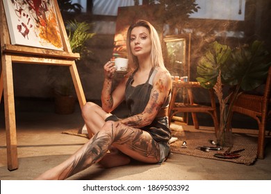 Posing in dark and atmospheric studio room beautiful tattooed and grimy woman holding coffee cup and looking at camera. - Powered by Shutterstock