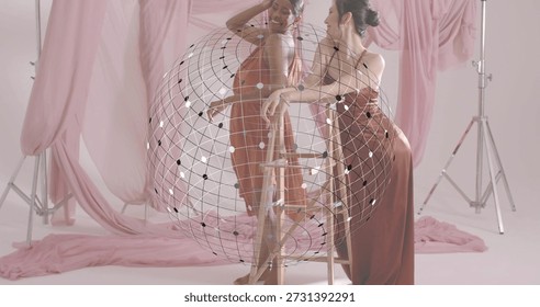 Posing two models in rust-satin leaning on wooden step-stool in pink studio with spherical grid. Duo, couture, elegance, fashion, pastel, soft, fabric