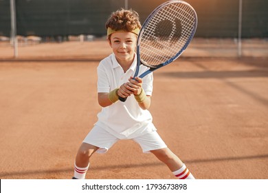 Positive active school aged boy with racket looking at camera while playing tennis on court