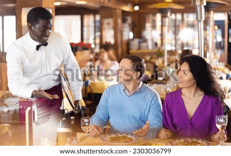Positive adult biracial couple talking to polite friendly African American waiter during dinner in cozy restaurant..